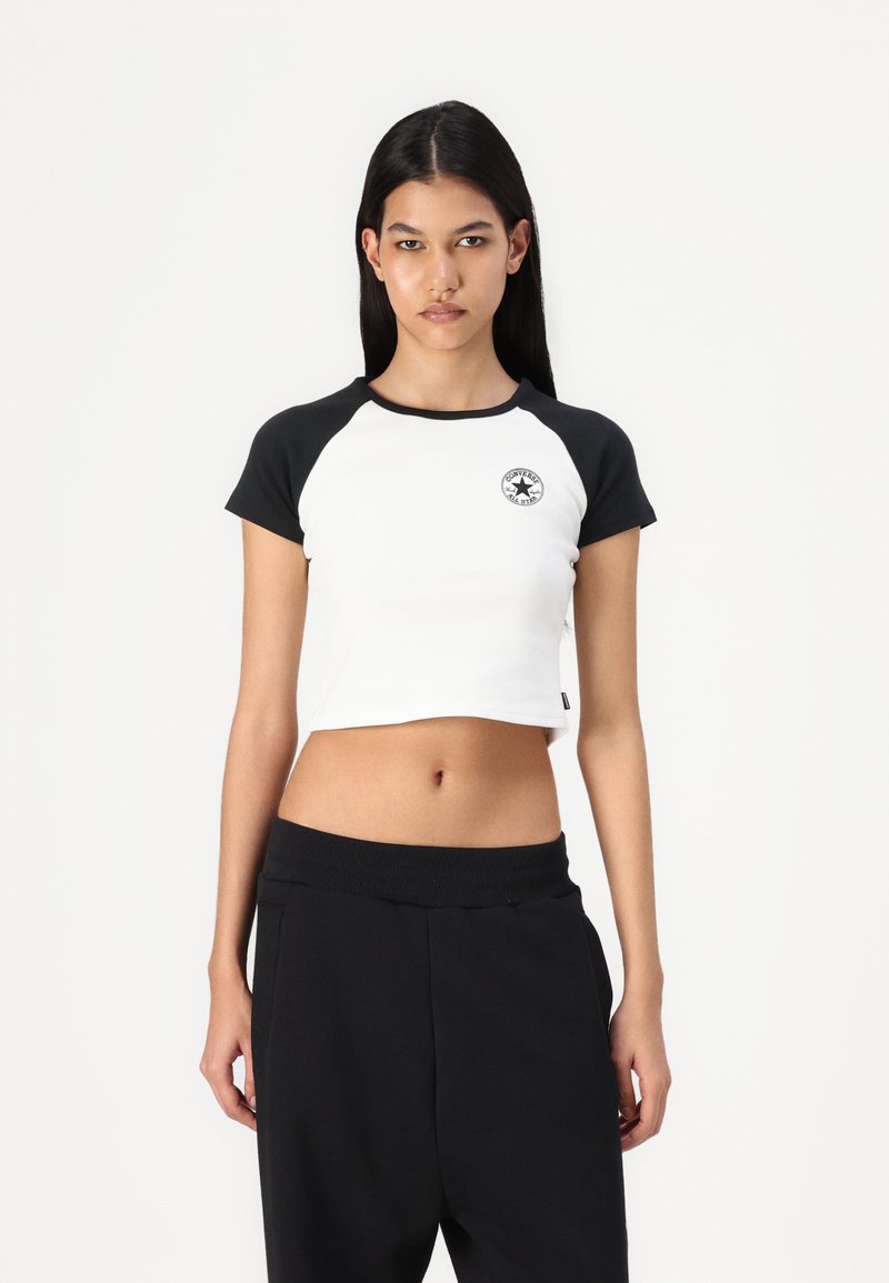 White cropped t-shirt with black raglan sleeves, featuring a circular logo on the chest. Worn with loose black pants. Smooth cotton texture.