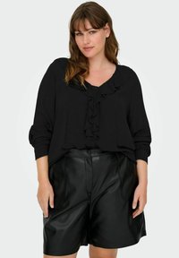 Sieviete ar vidēja garuma brūniem matiem, valkā melnu blūzi ar volāniem un garām piedurknēm, ielikta melnās ādas ceļgalu garuma šortos.