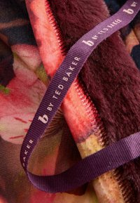 Mākslīgās kažokādas sega ar ziedu rakstu, kas izceļas ar bordo tekstūru un mīkstu virsmu. Ietver purpura krāsas zīmola etiķeti ar "Ted Baker".