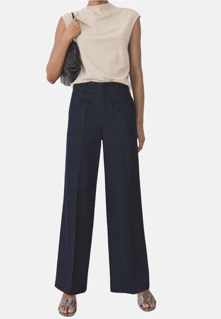Femme portant un haut beige sans manches, un pantalon large taille haute bleu marine, des sandales ouvertes en cuir façon serpent et portant un sac à épaule noir.