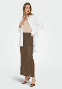 Witte knoop-overhemd over een beige tanktop, gecombineerd met een doorzichtige bruine rok met textuurpatronen en een zijsplit, gedragen met bruine hakken.