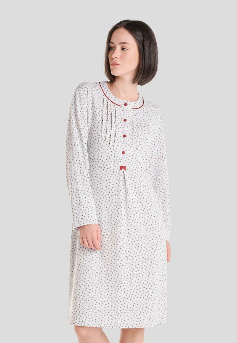 Camisa de noche blanca con pequeño estampado floral, mangas largas y cuello redondo. Presenta acentos rojos, botones y pliegues verticales en el escote.