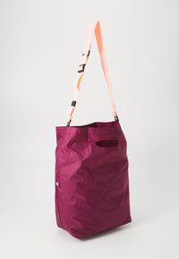 Bordo platnena torba s izrezom za ručku i podesivim svijetlopinkim remenom preko ramena na bijeloj pozadini.