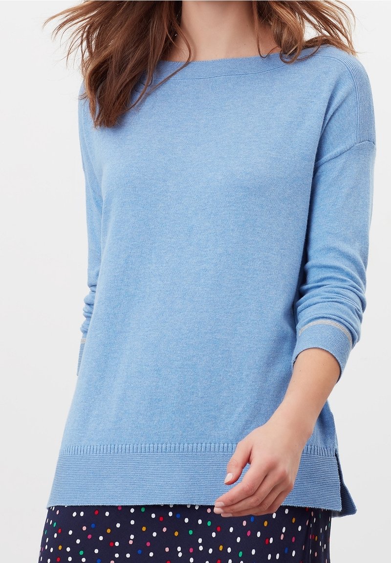 Femme portant un pull bleu clair à manches longues et une jupe foncée avec des pois colorés sur fond blanc.