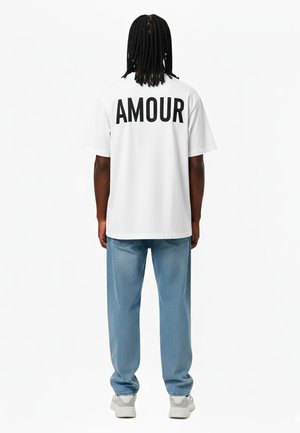 Witte t-shirt met "AMOUR" in het zwart gedrukt op de achterkant, gecombineerd met lichtblauwe jeans en witte sneakers. Casual, relaxte pasvorm.