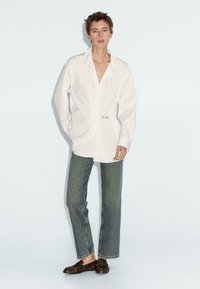 White oversized button-up shirt with a loose fit and pin detail, paired with straight-leg faded denim jeans and dark slip-on shoes.