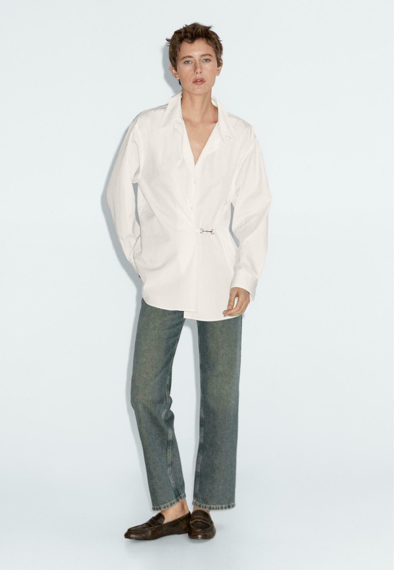 White oversized button-up shirt with a loose fit and pin detail, paired with straight-leg faded denim jeans and dark slip-on shoes.