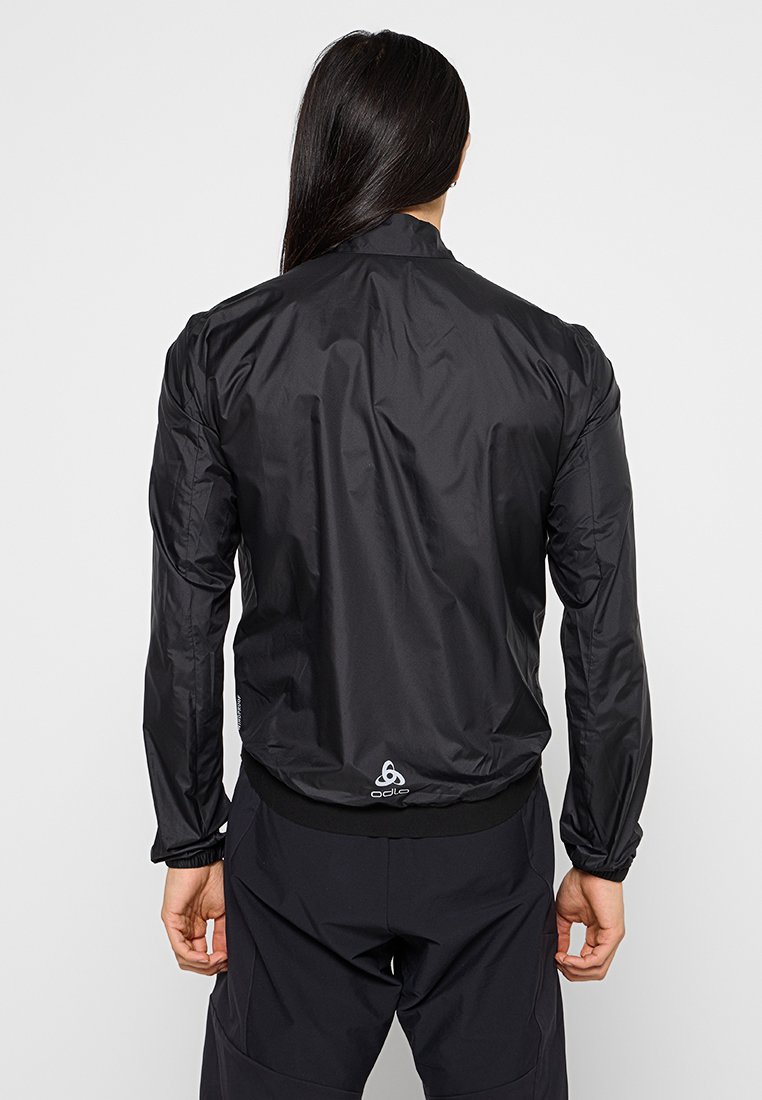 Mężczyzna z długimi ciemnymi włosami, ubrany w czarną lekką kurtkę i czarne spodnie, widoczny od tyłu na jednolitym jasnym tle.