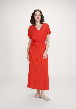 Femme aux cheveux bruns ondulés portant une robe portefeuille rouge à manches courtes avec une ceinture, assortie à des ballerines tissées beiges, debout sur un fond clair.