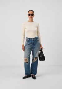 Beige ribbed long-sleeve top with frilled neckline, distressed blue baggy jeans with prominent rips, black accessories, and square-toed shoes.