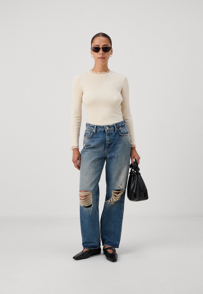 Haut beige à manches longues côtelé avec un col froncé, jean bleu baggy déchiré avec des déchirures visibles, accessoires noirs et chaussures à bout carré.
