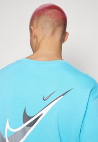Persona con pelo corto rosa que lleva una camisa de color azul claro con tres grandes logotipos de Nike superpuestos en la parte posterior.