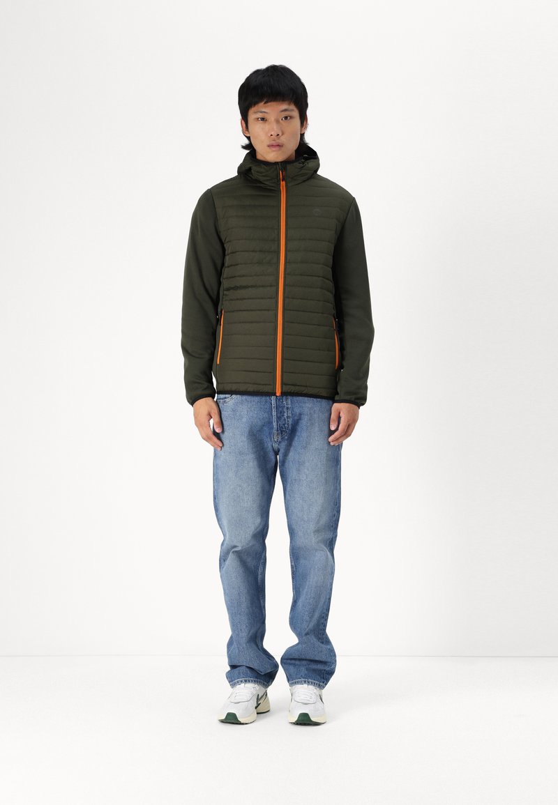 Olivgrüner Pufferjacke mit orangefarbenen Reißverschlüssen und Akzenten, ausgestattet mit Kapuze, gerippten Bündchen und leichter Bauweise. Kombiniert mit blauen Jeans.