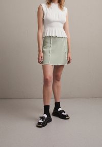 Bílý top bez rukávů s peplum střihem a texturovanou tkaninou, v kombinaci s světle zelenou sukní se zvlněným lemem a ozdobným lemem. Černé mokasíny s mašlemi.