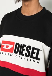 Svart sweatshirt med röda och vita horisontella ränder, stor "DIESEL"-logotyp i svart, mjuk textur och ribbad halsringning.