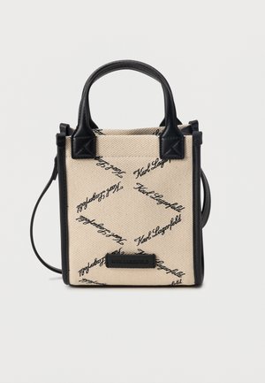 Mała beżowa torba na ramię z materiału, z czarnym napisem „Karl Lagerfeld”, czarnymi skórzanymi uchwytami, obszyciem i regulowanym paskiem na ramię.