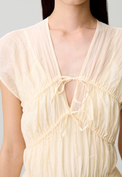 Donna che indossa un vestito in tessuto beige chiaro, arricciato, con maniche corte e due fiocchi annodati sulla profonda scollatura a V.