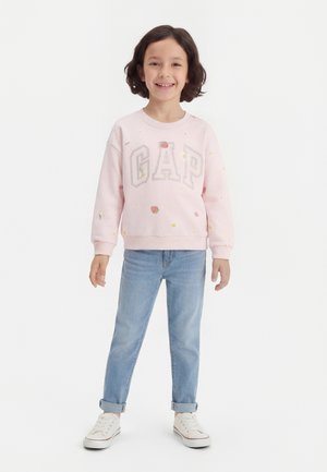 Smilende barn står iført en lyserød sweatshirt med teksten "GAP" og blomsterprint, lyseblå op rullede jeans og hvide sneakers.