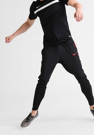Maglietta atletica nera con striscia bianca e pantaloni jogger neri con logo Nike rosso. Il modello indossa sneakers grigie, mostrando una posa dinamica.