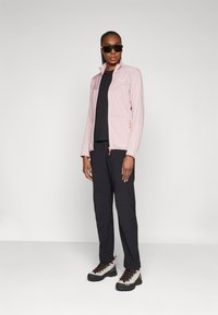 Lichtroze ritsjack met een verhoogde kraag, zwart t-shirt, zwarte broek en grijze laarzen met zwarte veters. Model staat voor een neutrale achtergrond.