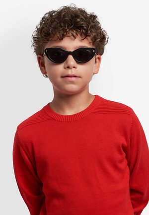 Bambino dai capelli ricci che indossa un maglione rosso e occhiali da sole neri a forma di occhi di gatto, in piedi davanti a uno sfondo grigio chiaro uniforme.