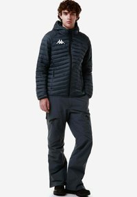 Giacca isolante scura con trapuntatura orizzontale e cappuccio, abbinata a pantaloni impermeabili grigi con tasche con zip e una vestibilità rilassata.
