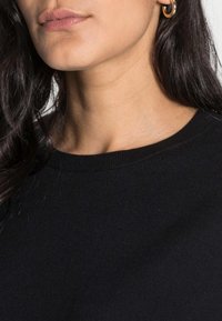 Sweat-shirt en coton noir avec un col rond et une texture côtelée. Gros plan montrant une peau lisse et une boucle d'oreille en or en forme d'anneau.