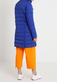 Abrigo puffer azul con costuras horizontales, mangas largas y cuello alto. Combinado con pantalones cortos de color naranja brillante y zapatillas blancas.