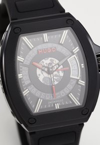 Black rubber watch with a square case. Transparent dial reveals intricate machinery. Silver and red accents highlight markers and hands.