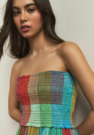 Young woman with long dark hair wearing a colorful strapless dress with a patchwork pattern against a plain light wall.