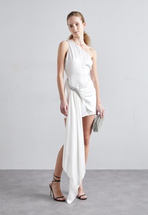 Witte satijnen jurk met asymmetrisch ontwerp, met gedrapeerde stof aan de ene kant, geaccentueerd door een zilveren versiering op de taille. Gecombineerd met een zilveren clutch.