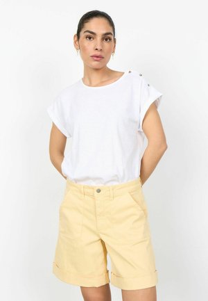 Woman wearing white short-sleeve top with button details and light yellow cuffed shorts, standing against white background.