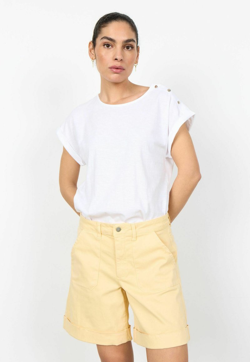 Femme portant un haut blanc à manches courtes avec des détails boutonnés et un short jaune clair avec revers, debout devant un fond blanc.
