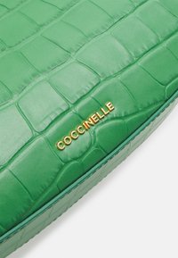 Zielona, teksturowana skóra z wytłoczonym złotym napisem "COCCINELLE", prawdopodobnie część torebki lub portfela.