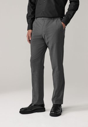 Mężczyzna w szarych, dopasowanych spodniach, czarnych eleganckich butach i czarnej koszuli zapinanej na guziki, z jedną ręką w kieszeni na jednolitym tle.