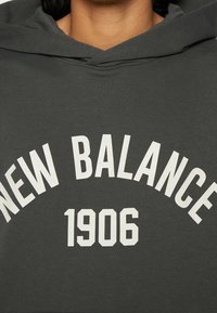 Felpa con cappuccio grigia con una grafica bianca stampata che riporta la scritta "NEW BALANCE" e "1906" in lettere maiuscole e in grassetto sulla parte anteriore.