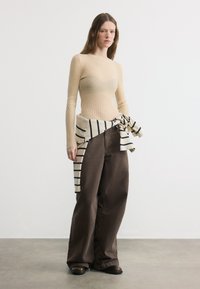 Femme portant un haut beige à manches longues côtelées, un pantalon large marron, et un pull rayé noué autour de la taille, debout sur un sol gris.