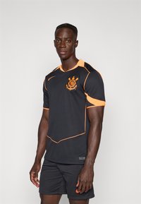 Svart atletisk skjorte med oransje detaljer og krage. Designet inkluderer en logo på brystet og mesh-tekstur, kombinert med svarte shorts.