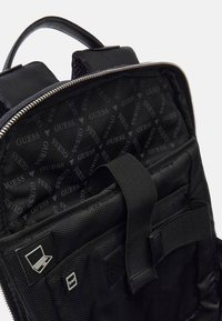 Black backpack interior with a textured fabric lining featuring a repeated "GUESS" logo pattern, mesh pocket, and adjustable strap details.