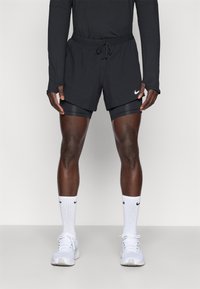 Schwarze Sportshorts mit verstellbarem Bund, über schwarzen Kompressionsshorts getragen. Weißer Nike-Swoosh an der Seite, kombiniert mit weißen Sneakers.