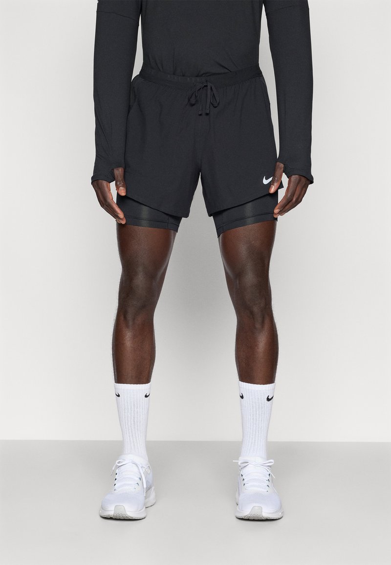 Schwarze Sportshorts mit verstellbarem Bund, über schwarzen Kompressionsshorts getragen. Weißer Nike-Swoosh an der Seite, kombiniert mit weißen Sneakers.