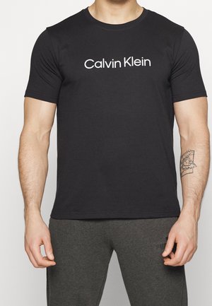 Muž nosící černé tričko Calvin Klein a tmavě šedé tepláky s tetováním na pravém předloktí.
