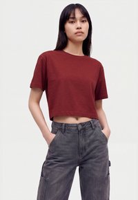 Giovane donna con capelli neri lisci indossa una t-shirt rossa corta e pantaloni cargo neri scoloriti a vita alta, con le mani nelle tasche.