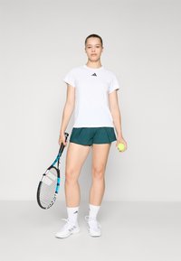 T-shirt de sport blanc avec logo noir, shorts verts, raquette de tennis à la main, balle de tennis jaune, chaussettes blanches et chaussures. Design simple.