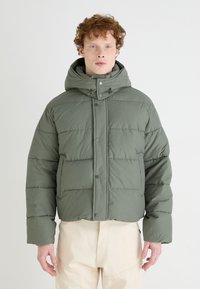 Green puffer jacket with a hood, featuring a boxy design, snap button closure, and quilted texture. Worn over beige trousers.
