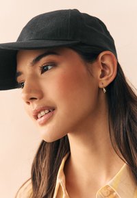 Sort kasket med buet skygge, båret med en lysegul skjorte med krave. Har en glat tekstur og minimalistisk design.