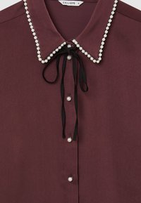 Blusa bordeaux con colletto rifinito in perle, cravatta nera al collo e bottoni in perla lungo la parte anteriore.