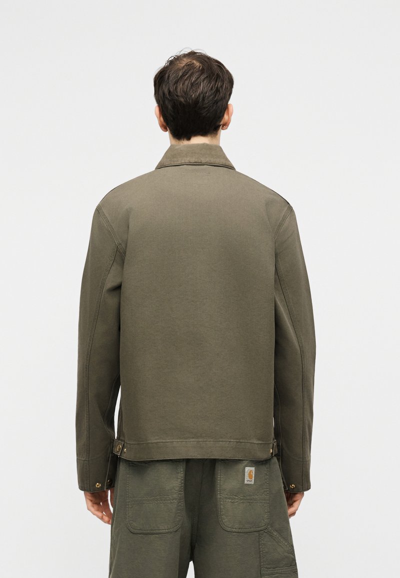 Uomo con giacca verde oliva e pantaloni cargo abbinati, visto di spalle su uno sfondo chiaro e uniforme.