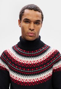 Homme avec les cheveux courts et bouclés portant un pull à col roulé noir avec un motif tricoté rouge, blanc et gris.