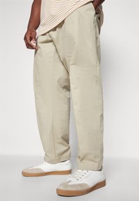 Pantaloni beige chiaro in cotone con vestibilità rilassata, tasche laterali e una texture leggermente stropicciata. Abbinati a sneakers bianche e marroni.
